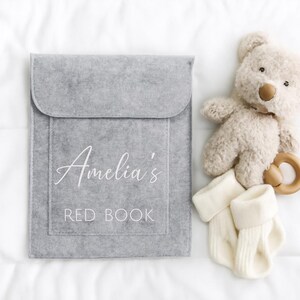 Op de afbeelding: Een grijze vilten boekomslag met de tekst "Amelia's RED BOOK" in witte letters. Naast de boekomslag liggen een beige teddybeer, een houten bijtring en een paar witte babysokjes. De items liggen op een witte ondergrond.