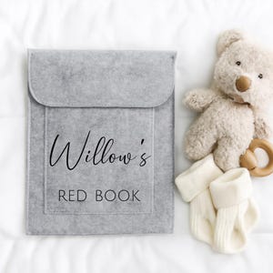 Op de afbeelding: Een grijze vilten boekomslag met de tekst "Willow's RED BOOK" in zwart schrift. Daarnaast een beige teddybeer, een houten bijtring en een paar witte babysokjes. De items zijn gerangschikt op een wit oppervlak.