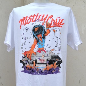 以下が含まれることがあります： 白いTシャツ。オレンジ色の「Mötley Crüe」のバンド名と、スタジアムを突き破るモンスターのカラフルなグラフィックが特徴です。紫とオレンジの色合いのコンサートシーンが含まれています。