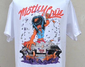 May include: White t-shirt featuring a colorful graphic of a monster breaking through a stadium, with the band name "Mötley Crüe" in orange. The design includes a concert scene with purple and orange hues.