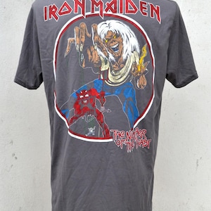 Puede incluir: Camiseta gris oscuro con un gráfico de una figura esquelética y un diablo rojo. El texto "IRON MAIDEN" está arqueado sobre la imagen, con "THE NUMBER OF THE BEAST" debajo. El diseño está en un círculo rojo y blanco.