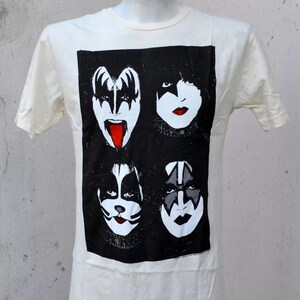 May include: White t-shirt with a black and white image of the four members of the rock band KISS. The image shows the band members' faces with their signature makeup.