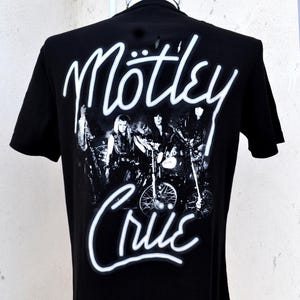 May include: Black t-shirt featuring a black and white image of a band on motorcycles. The band's name, "Mötley Crüe", is written in white, stylized script above and below the image. The shirt is displayed on a mannequin.