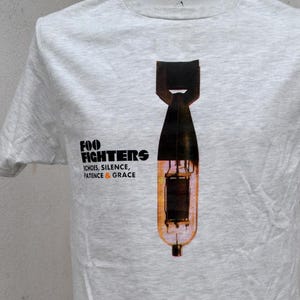 May include: A light gray t-shirt featuring a graphic of a bomb with a black top and a brown body. The text "FOO FIGHTERS" is printed above the bomb, with "ECHOES, SILENCE, PATIENCE & GRACE" below. The shirt has short sleeves.