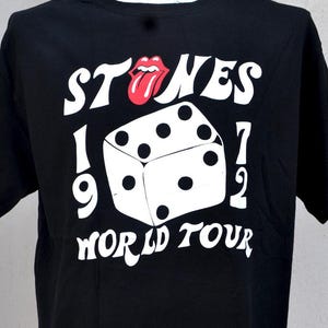 May include: Black t-shirt with a white graphic featuring the word "STONES", a red tongue logo, a die, and the text "1972 WORLD TOUR". The shirt is displayed on a mannequin.