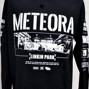 May include: A black long-sleeve shirt featuring white graphic text and imagery. The front displays the word METEORA above a rectangular black and white scene, the text LINKIN PARK, a tracklist, the year 2003, and additional graphic symbols.