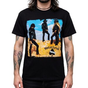Può includere: T-shirt nera con una grafica della band Motörhead. Il design include il nome della band, un simbolo di picche e un'immagine dei membri della band. L'immagine ha uno sfondo desertico con un cielo blu.