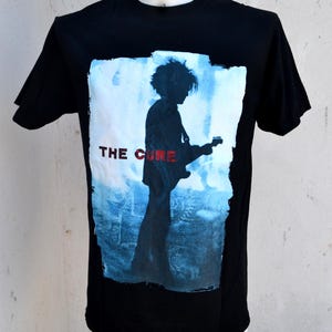 Może przedstawiać: Czarny t-shirt z sylwetką gitarzysty na niebiesko-białym tle. Czerwony napis "THE CURE" jest nadrukowany z przodu. Koszulka ma krótkie rękawy i okrągły dekolt. Odzież o tematyce muzycznej.