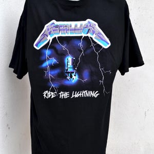 May include: Black t-shirt featuring the band name "Metallica" in a blue and purple design above a graphic of a chair and lightning bolts. The text "Ride the Lightning" is below the graphic. The shirt is displayed on a mannequin.