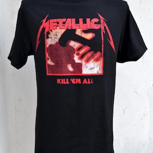 May include: Black t-shirt featuring the band name "METALLICA" in red, with a red and orange graphic of a hand. The text "KILL 'EM ALL" is printed below the graphic. The shirt is displayed on a mannequin.