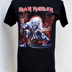 Puede incluir: Camiseta negra con el nombre de la banda "Iron Maiden" en letras rojas sobre un gráfico de una figura esquelética con piel azul, pelo blanco y detalles rojos. La figura sostiene palos y está rodeada de llamas rojas y naranjas.