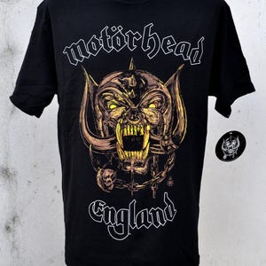 May include: Black t-shirt featuring the band name "Motörhead" and a detailed illustration of a snarling, horned creature with chains and skulls. The word "England" is also displayed. A small circular logo is visible.