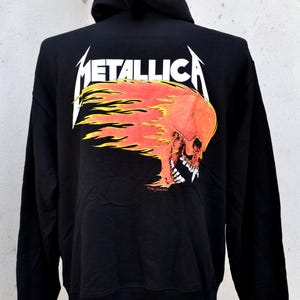 Può includere: Felpa con cappuccio nera con il nome della band "METALLICA" in bianco, sopra una grafica di teschio fiammeggiante in rosso e arancione. La felpa con cappuccio ha un cappuccio con coulisse e maniche lunghe. Un classico capo d'abbigliamento rock.
