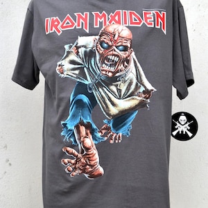 May include: A dark grey t-shirt with the red text "IRON MAIDEN". The graphic shows a zombie-like figure with blue eyes, torn clothing, and a chain. The figure is in a dynamic pose. A small logo is visible on the right side of the t-shirt.