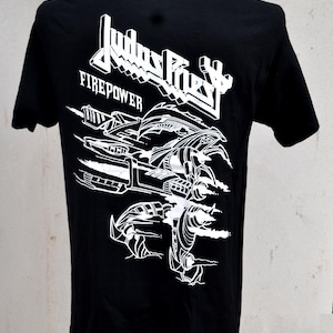 Puede incluir: Camiseta negra con un diseño gráfico blanco y texto. El diseño incluye el nombre de la banda "Judas Priest" y la palabra "Firepower" con una ilustración de un vehículo futurista. La camiseta se muestra en un maniquí.