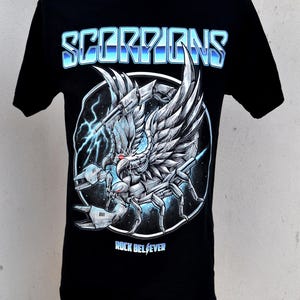 May include: Black t-shirt featuring the band name "SCORPIONS" in a blue gradient font above a detailed illustration of a scorpion with wings and lightning bolts. The text "ROCK BELIEVER" is below the graphic.