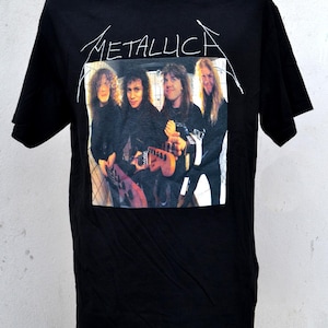Op de afbeelding: Zwart T-shirt met de naam van de band Metallica in witte letters. Onder het logo staat een rechthoekige afbeelding van de bandleden. De bandleden dragen zwarte kleding en houden instrumenten vast.