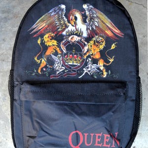 Puede incluir: Mochila negra con el escudo de Queen, que incluye una corona, un águila y leones en dorado, rojo y blanco. La palabra "QUEEN" está impresa en rojo en el bolsillo delantero. Tiene un bolsillo lateral de malla.