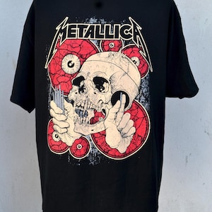 Puede incluir: Camiseta negra con un diseño gráfico de una calavera, ojos rojos y el nombre de la banda "METALLICA" en blanco. El diseño tiene un aspecto envejecido. La camiseta es de cuello redondo.
