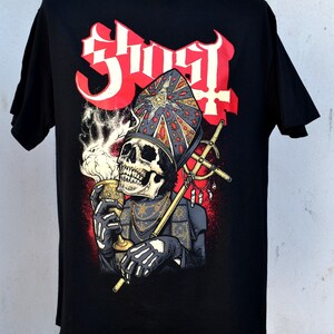Puede incluir: Camiseta negra con un diseño de una figura esquelética con una mitra de obispo, sosteniendo un cáliz y un báculo. La palabra "Ghost" está en letras rojas estilizadas. El diseño incluye detalles en rojo y una cruz.