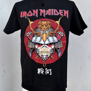 May include: Black t-shirt featuring the band name "Iron Maiden" in red and a graphic of a samurai warrior's mask. The design includes red, gold, and white accents, with Japanese characters below the graphic.
