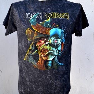 Peut inclure: T-shirt noir avec un graphisme de visage cyborg et le nom du groupe "Iron Maiden" en jaune et blanc. Le design présente une illustration détaillée et colorée avec un aspect vieilli et texturé.