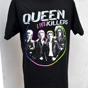 Puede incluir: Camiseta negra con la inscripción "QUEEN LIVE KILLERS" en blanco y rosa. El diseño incluye una imagen en blanco y negro de la banda Queen dentro de un círculo de colores del arcoíris. La camiseta se muestra en un maniquí.