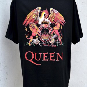 Può includere: T-shirt nera con un design a stemma colorato raffigurante una fenice, leoni e una corona. La parola "QUEEN" è stampata in rosso sotto. La maglietta è esposta su un manichino.