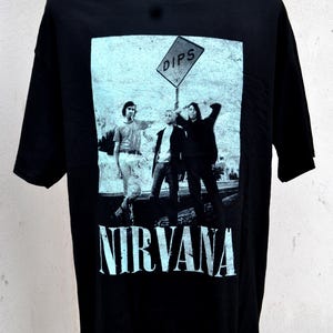 May include: Black t-shirt featuring a graphic of the band Nirvana. The graphic is a black and white image of three people standing near a road sign that says "DIPS". The band's name, "NIRVANA", is printed in large, light blue letters below the image.