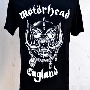 Może przedstawiać: Czarny t-shirt z białym nadrukiem czaszki z rogami, łańcuchami i napisem "motörhead England". Projekt jest wyśrodkowany na przodzie koszulki, z nazwą zespołu nad czaszką.