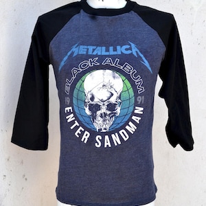 Op de afbeelding: Een raglan-stijl T-shirt met een zwart en blauw ontwerp. Het shirt heeft de bandnaam "Metallica" in blauw, boven een schedelafbeelding met de tekst "Black Album", "Enter Sandman" en "91".