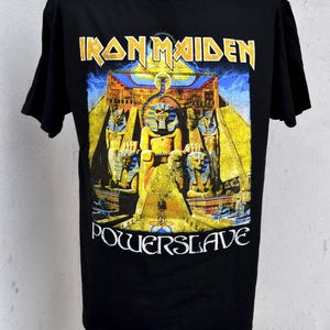 Op de afbeelding: Zwart T-shirt met de bandnaam "Iron Maiden" in geel en een Egyptisch thema-ontwerp met het woord "Powerslave" in wit. Het ontwerp bevat gele, blauwe en oranje tinten.