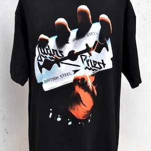 Op de afbeelding: Zwart T-shirt met een afbeelding van een scheermes, de woorden "British Steel" en "Judas Priest". Het ontwerp bevat een oranje hand en een zwarte armband met zilveren studs. Het shirt wordt op een mannequin getoond.