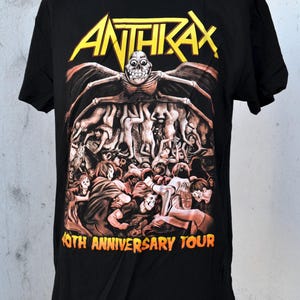 Puede incluir: Camiseta negra con el nombre de la banda "ANTHRAX" en letras amarillas. El gráfico representa una figura esquelética con los brazos extendidos y una multitud de personas. El texto "40TH ANNIVERSARY TOUR" está impreso en naranja.