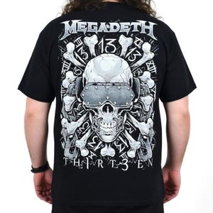 Può includere: Una t-shirt nera a maniche corte con una stampa grafica di un teschio che indossa occhiali protettivi metallici, circondato da ossa incrociate e dal testo "MEGADETH" in alto e "THIRTEEN" in basso, con il numero "13" ripetuto sullo sfondo.