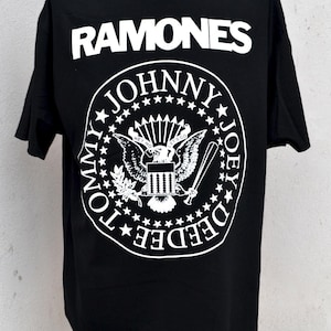 Puede incluir: Camiseta negra con la palabra "RAMONES" en blanco sobre un emblema circular. El emblema presenta los nombres de los miembros de la banda y un diseño de águila. Una prenda clásica con temática musical.
