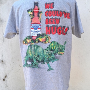以下が含まれることがあります： ヘザーグレーの半袖Tシャツ。バドワイザーのビール瓶、サングラスをかけた3匹のカエル、2匹のカメレオンのグラフィックプリントが施されています。右側には「WE COULD'VE BEEN HUGE!」という文字が大きな赤い文字でプリントされています。