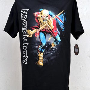 Peut inclure: T-shirt noir avec un motif d'une figure squelettique en veste rouge, pantalon bleu et bottes dorées, tenant un drapeau. Le texte "IRON MAIDEN" est imprimé verticalement sur le côté gauche du t-shirt.