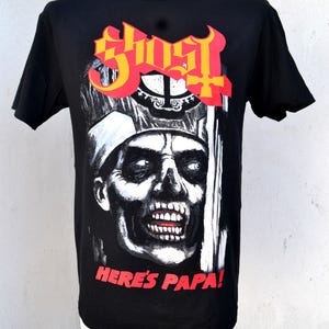 May include: Black t-shirt featuring a graphic of a skeletal figure with the text "GHOST" in red and yellow, and the phrase "HERE'S PAPA!" in red. The design is a detailed illustration.