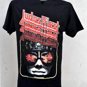 May include: Black t-shirt with the band name "Judas Priest" in red and white. The shirt features a graphic of a face wearing sunglasses with red and orange reflections. The graphic is framed by a yellow border.