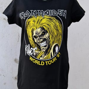 Puede incluir: Camiseta negra con un gráfico amarillo y blanco de una figura con cara de calavera y pelo amarillo. El texto "IRON MAIDEN" está encima del gráfico, y "Killer World Tour '81" debajo.