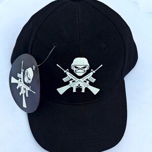 May include: Black baseball cap with a white graphic of a skull wearing a helmet and crossed rifles. A matching tag is attached to the cap. The cap is made of a durable fabric and has a curved brim.