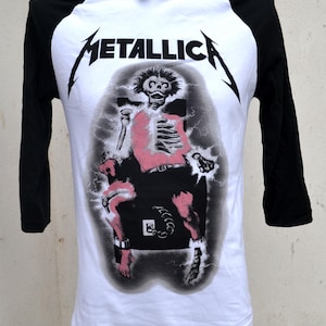 Op de afbeelding: Wit en zwart raglan mouwen T-shirt met de bandnaam "METALLICA" in zwart. De afbeelding toont een roze en zwart skelet in een elektrische stoel, met een grijze achtergrond en bliksem.
