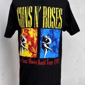Puede incluir: Camiseta negra con el logo de Guns N' Roses en amarillo y el texto "Use Your Illusion World Tour 1992". El diseño incluye dos cuadrados con imágenes de una persona sobre fondos rojos y azules.