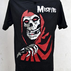 May include: Black t-shirt with a skull graphic, red eyes, and a red hooded cloak. The word "Misfits" is printed in white. The shirt is displayed on a mannequin. The design is a classic punk rock image.