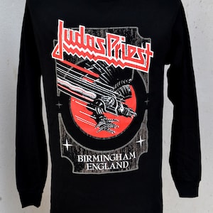 May include: Black long-sleeve shirt featuring the band name "Judas Priest" in red and white, with a graphic of a winged creature and the text "Birmingham England". The shirt is displayed on a mannequin.