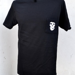 May include: Black t-shirt with a white skull graphic on the pocket. The short-sleeved shirt is displayed on a mannequin. The skull graphic is a stylized, white design.