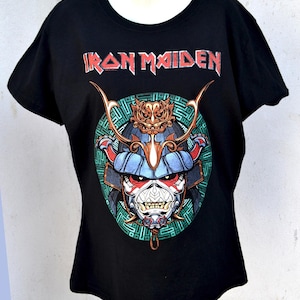 May include: Black t-shirt featuring the band name "Iron Maiden" in red lettering above a graphic of a samurai warrior's face. The warrior has a blue helmet, red face, and gold accents, set against a green and blue circular background.