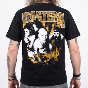 Può includere: T-shirt nera con la band Pantera. Il design presenta una foto di gruppo dei membri della band in bianco e giallo, con il nome della band 'Pantera' in giallo testurizzato in alto.
