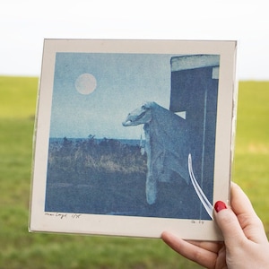 Op de afbeelding: Een blauwe en witte print van een landschap met een grote maan aan de hemel. De print is ingelijst en heeft de tekst "man Langel" en "1/97" in de linkerbenedenhoek.
